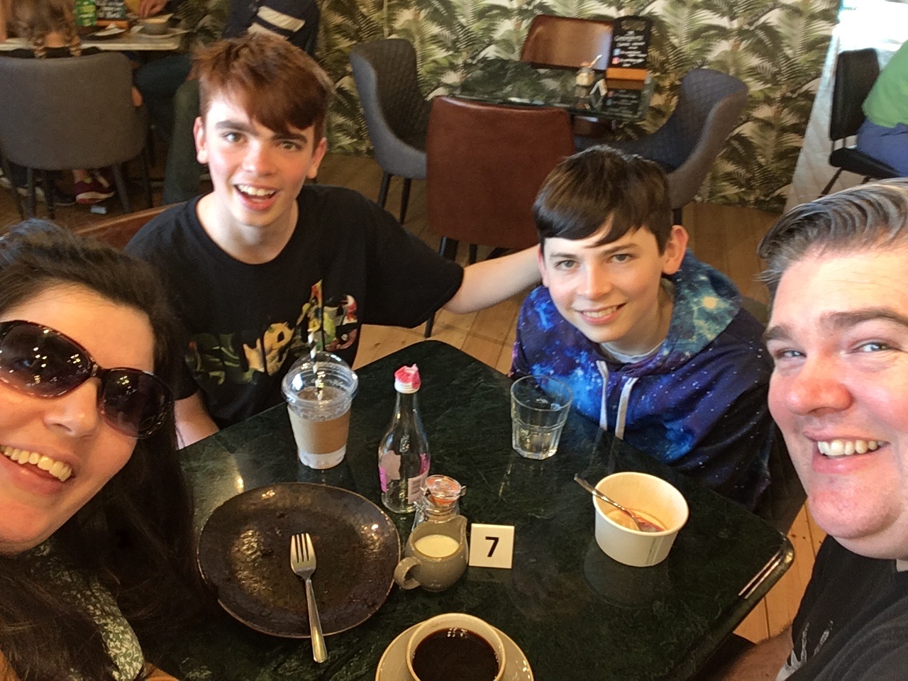A selfie of Joel, Myself and our boys round the table in a coffee shop. I’m wearing red sunglasses and you can only see mine and Joel's face. Youngest is warring a black graphic T-shirt and Eldest is wearing a blue galaxy scene hoodie. 